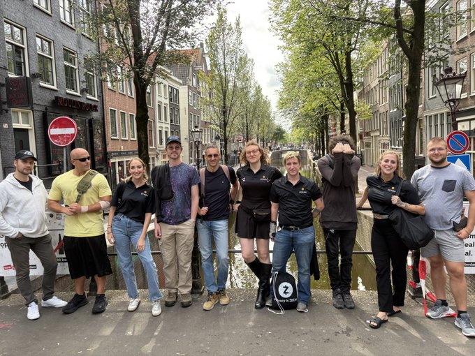 A group of nine tour participants poses on a bridge in Amsterdam's oldest district, with one person holding a "Zcash" branded bag featuring the number "2" during the Red Light Cypherpunks Walking Tour exploring privacy and history.