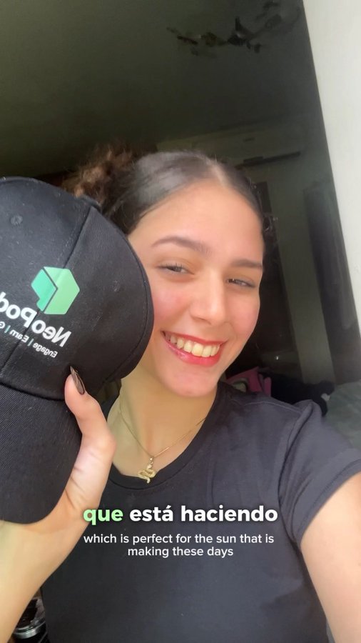 A smiling young woman holds up a black cap with a green 'NeoDob' logo, with subtitles reading 'que está haciendo' and 'which is perfect for the sun that is making these days'.