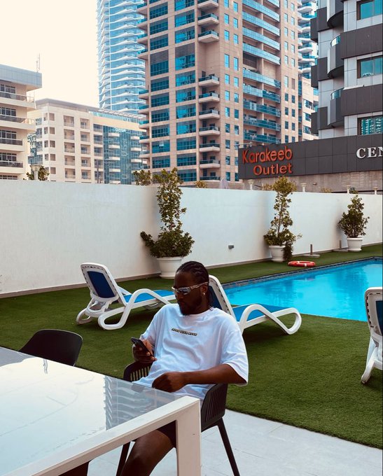 A man in a white t-shirt and sunglasses sitting at an outdoor table using his phone by a rooftop pool, with modern high-rise buildings and a 'Karakeeb Outlet' sign in the background.