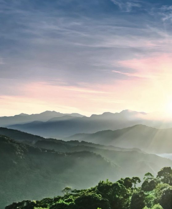 A scenic mountain landscape at sunrise or sunset, with layers of misty mountains, sun rays breaking through the sky, and green vegetation in the foreground.