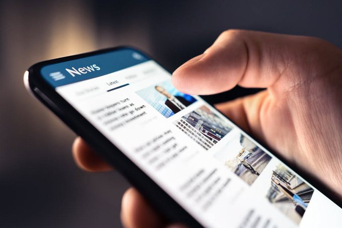 A close-up shows a hand holding a smartphone displaying a 'News' application interface with various article thumbnails and text.