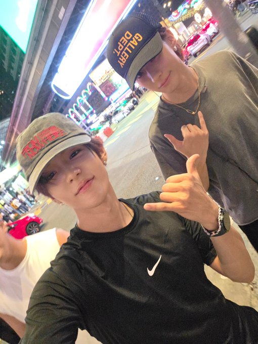 Two young men pose for a nighttime street selfie in a city with neon lights, both making hand gestures, with the front person wearing a "Galaxy Dept." cap and a black Nike t-shirt, and the back person wearing a cap with red text and a gray shirt.