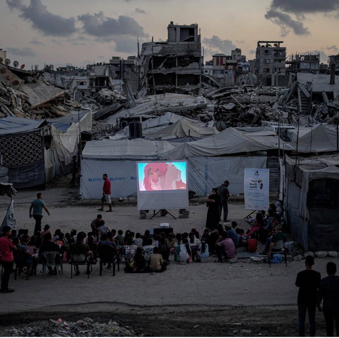 Children sitting in plastic chairs watching an animated film projected onto a screen in a tent encampment, set against a backdrop of heavily bombed and destroyed buildings at dusk.
