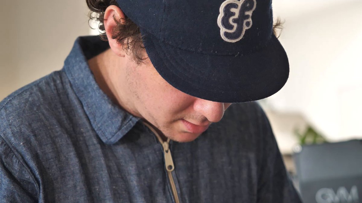 The image features a man wearing a blue baseball cap with an EFDOT studio logo on it. The man appears to be looking down at something, possibly a book or a piece of paper. The background shows a white wall, which contrasts nicely with the dark blue color of the cap and adds depth to the scene. The caption above the image reads "Just a couple hours away from one of the most compelling collection drops we’ve seen in a while, GLiFs by @EfdotStudio". This suggests that there is an upcoming event or release related to the EFDOT studio's collection drop, and the man might be preparing for it.