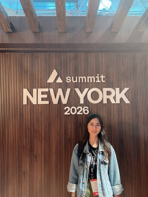 A woman with glasses wearing a denim jacket poses in front of a wooden slatted wall displaying 'summit NEW YORK 2026' in white lettering.