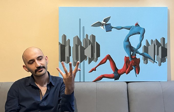 A bald man in a dark shirt sits on a gray couch, gesturing with his hand while speaking, with a large painting behind him depicting two superhero figures—one in blue and one in red—engaged in a dramatic confrontation against a geometric cityscape background, matching the post's "Me and my art" caption.
