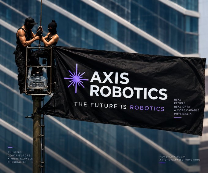 Two masked figures stand on a platform rig attaching a large black banner that reads "AXIS ROBOTICS - THE FUTURE IS ROBOTICS" with tagline "Real People, Real Data, A More Capable Physical AI," along with corner text "Builders Contributors A More Capable Physical AI" and "More Data Today A More Capable Tomorrow."

The image visually reinforces the post's message by presenting Axis Robotics' branding through a guerrilla-style banner installation, framing the company's mission of crowdsourced robot teleoperation data as a bold, almost revolutionary act that transforms human contributors into builders of Physical AI.
