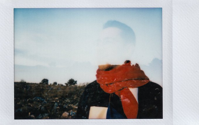 A faded Polaroid-style photograph showing a person wearing a red scarf in an outdoor setting with sky and landscape visible.