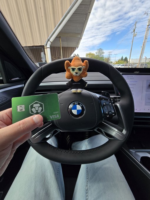 A first-person view from the driver's seat of a BMW, showing a hand holding a green Crypto.com Visa debit card, with a small stuffed lion toy attached to the steering wheel.