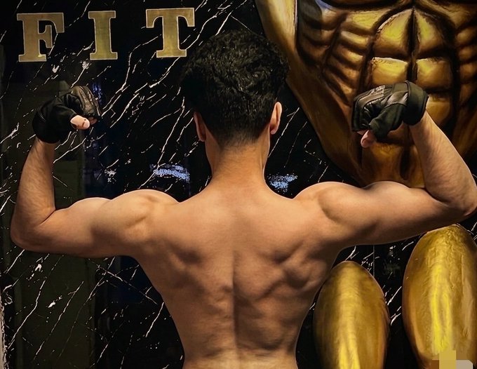 A shirtless man flexes his back muscles and biceps while wearing MMA gloves in a gym, with a "FIT" logo visible on the dark marble wall behind him, perfectly matching the "GM CT Happy Saturday everyone Gym" post about a Saturday gym session.