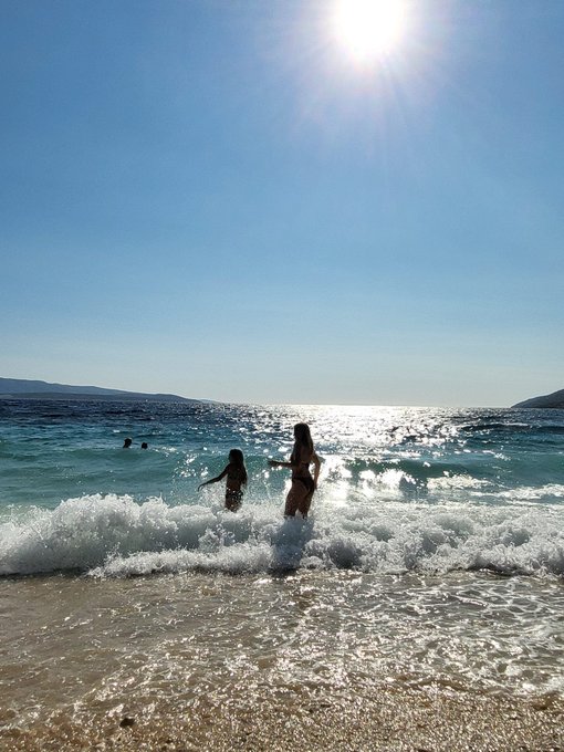 A scenic beach view with two people playing in the surf under a bright, sunny sky.