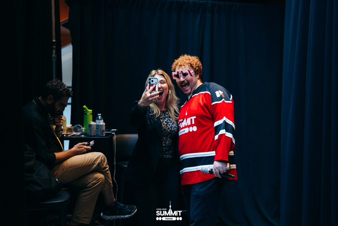 A photo from 'The Summit' event showing a woman taking a selfie with a man in a red hockey jersey and novelty star-shaped glasses, while another person sits in the background.
