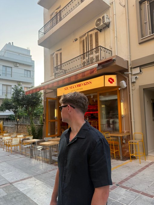 A man wearing sunglasses and a dark shirt stands in front of a restaurant with a sign reading 'SIX SECOND KISS' featuring a lips logo.