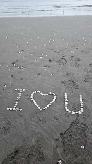 A black and white beach photograph featuring the words 'I [heart] U' spelled out with white seashells on the dark sand, with footprints leading toward the ocean waves.