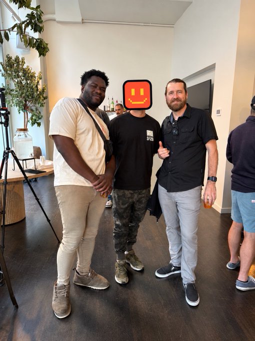 Three men pose together indoors; the central figure wears a black t-shirt with his face obscured by a yellow smiley-face emoji, flanked by two men who appear to be of different races.