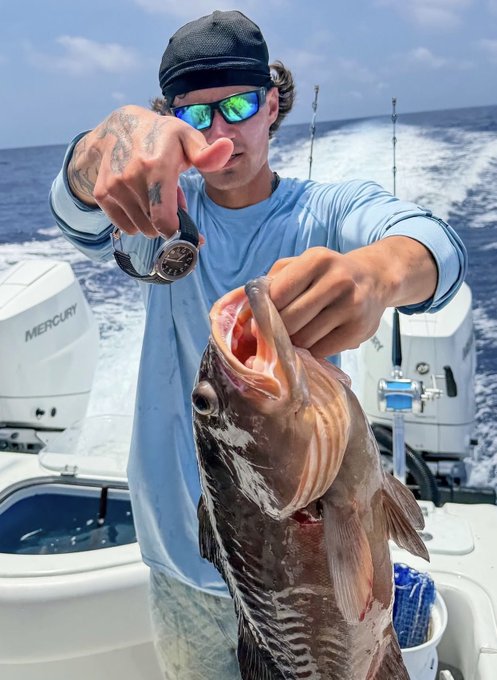 A fisherman wearing mirrored shades proudly shows off a hefty catch on a boat powered by a "Mercury" outboard engine, with the sporty ocean backdrop complementing the rugged vibe of the image.