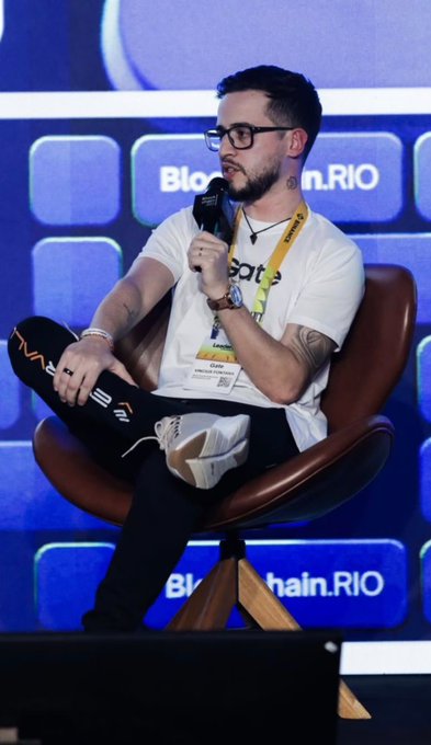 A young man with glasses and a beard sits on a brown leather chair, speaking into a microphone while wearing a white t-shirt, ripped black jeans, and a "Block Chain.RIO" lanyard with an event pass. The backdrop displays the "Block Chain.RIO" logo, tying the scene to the accompanying post celebrating his milestone of speaking on the Main Stage of the largest Web3 event in LATAM.