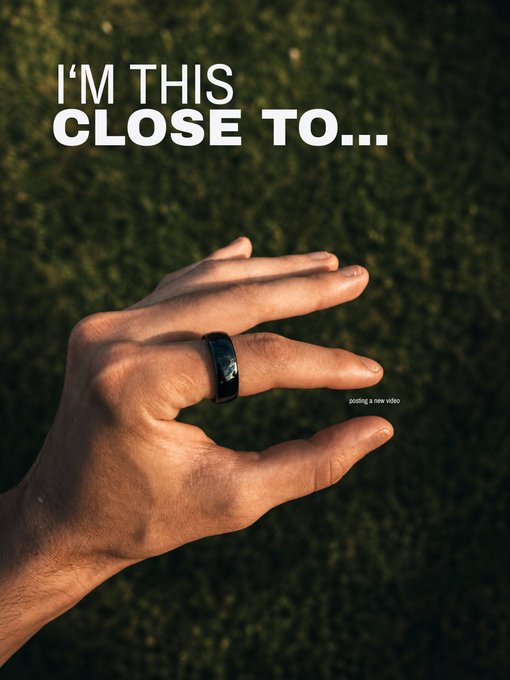 A hand is held up against a grassy background displaying a black ring on the middle finger, with text reading 'I'M THIS CLOSE TO...' and 'posting a new video'.
