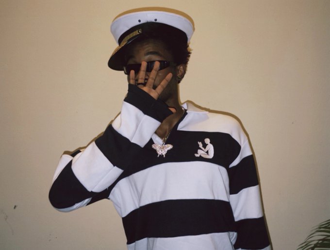 A man wearing a captain's hat, sunglasses, and a black and white striped long-sleeve shirt poses with his hand near his face, displaying a jeweled pendant necklace.