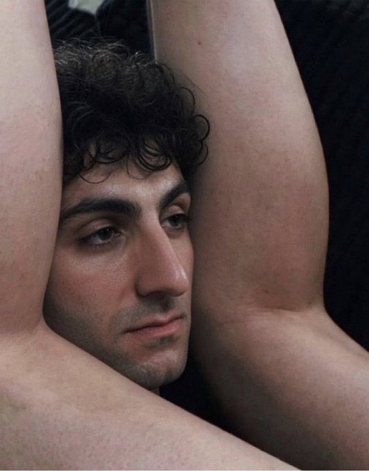 A shirtless man with dark curly hair looks off to the side with a sultry expression, his arms raised and framing his face—evoking the dramatic, brooding intensity often associated with stock chart "GM" (good morning) posts seeking attention for Solana ($SOL).