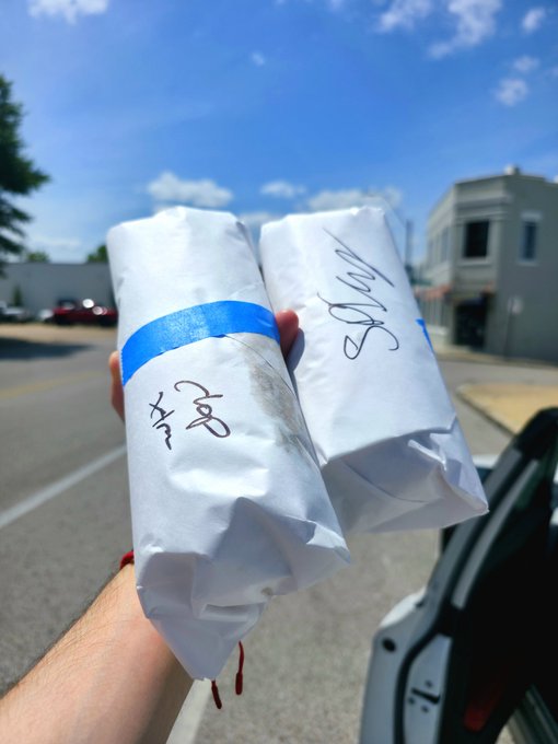 The image shows a hand holding two wrapped sandwiches, presumably for someone else or as a treat for themselves. The sandwiches are wrapped in white paper with blue tape, which adds a pop of color to the scene. The background features a city street lined with buildings, indicating that this is likely taking place in an urban setting. The post appears to be promoting a sandwich shop or a similar establishment and possibly encouraging people to buy lunch for themselves or others as a gesture of kindness or support.