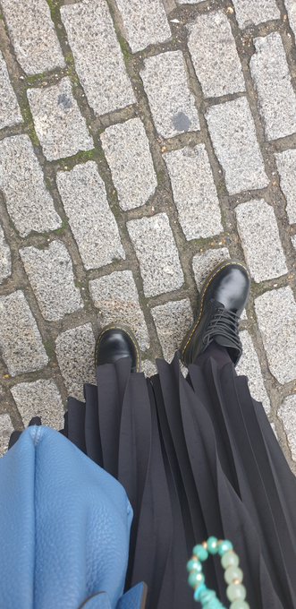 An overhead photo showing a person's feet in black Dr. Martens-style boots on a cobblestone surface, wearing a black pleated skirt and carrying a blue bag with a turquoise beaded bracelet visible.