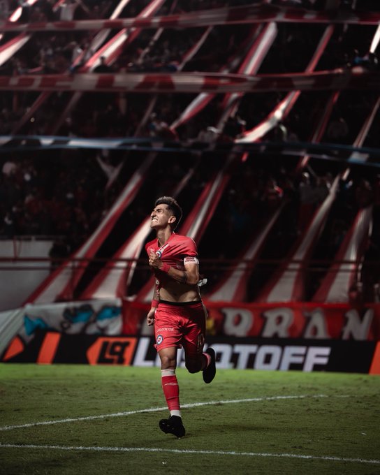 A jubilant soccer player in a red #10 jersey celebrates a goal on the pitch, as fans wave red and white striped banners behind him and advertising boards display "PLAYOFF" text, suggesting the moment ties into the excitement around securing Lescano's transfer.