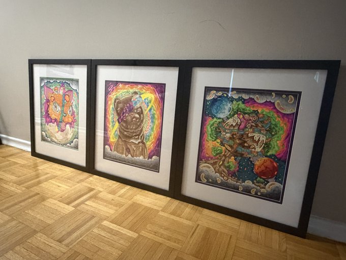 Three framed colorful artworks are displayed leaning against a beige wall on a wooden parquet floor, featuring vibrant psychedelic and cosmic imagery.