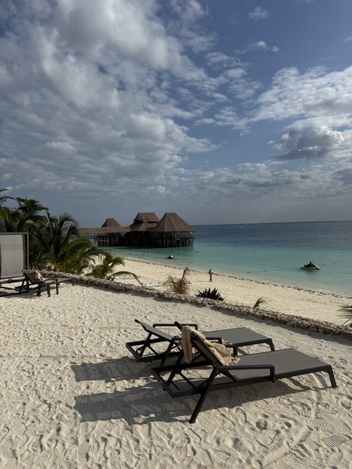 A serene beach scene with lounge chairs on white sand, turquoise water, and overwater bungalows in the distance, evoking a perfect "Good Morning" paradise. The image relates to the "GM" (Good Morning) post by showcasing a tranquil morning view at what appears to be a tropical resort.