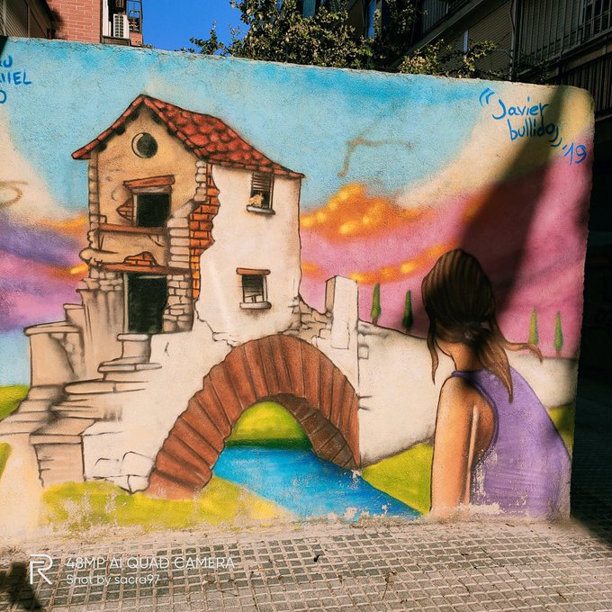 Street mural signed 'Javier bullido 19' depicting a woman in a purple dress looking toward a rustic house with a stone bridge and sunset sky.