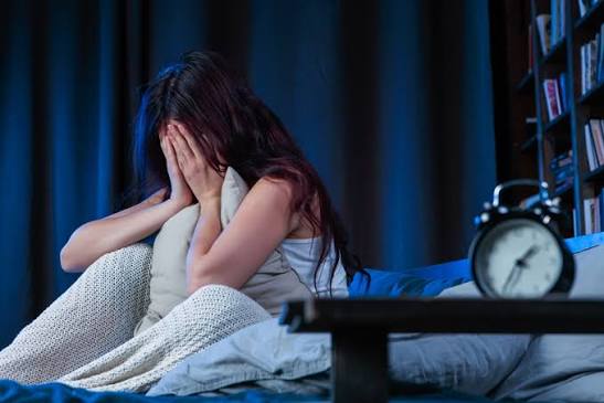 A young woman with messy dark hair sits on a bed at night, covering her face with her hands next to an analog alarm clock on a nightstand.