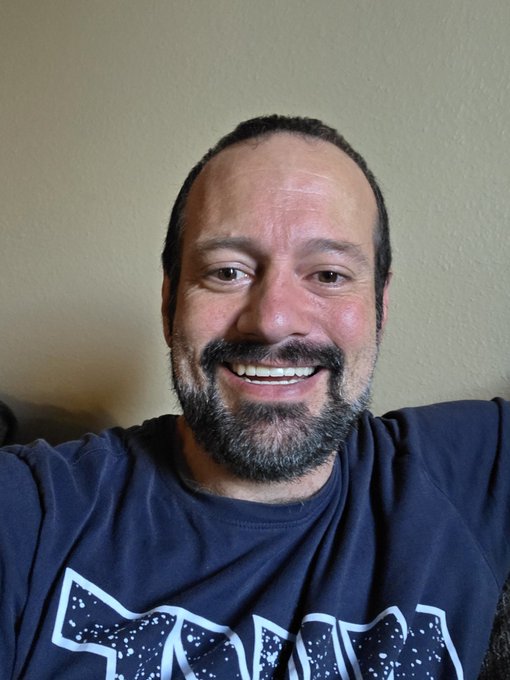 A bearded man smiles brightly, showing off his fixed tooth, wearing a dark blue graphic t-shirt, thanking the ICP and CKB communities for their support that allowed him to get his dental work done and return to making content.