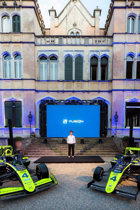 A man stands on a stage in front of a large screen displaying 'LIGIER FUSION,' flanked by two green and black racing cars in a courtyard.
