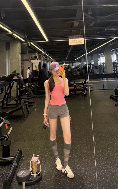 A woman in a pink tank top, gray shorts, and knee-high socks taking a mirror selfie in a gym, with workout equipment and other people visible in the background.
