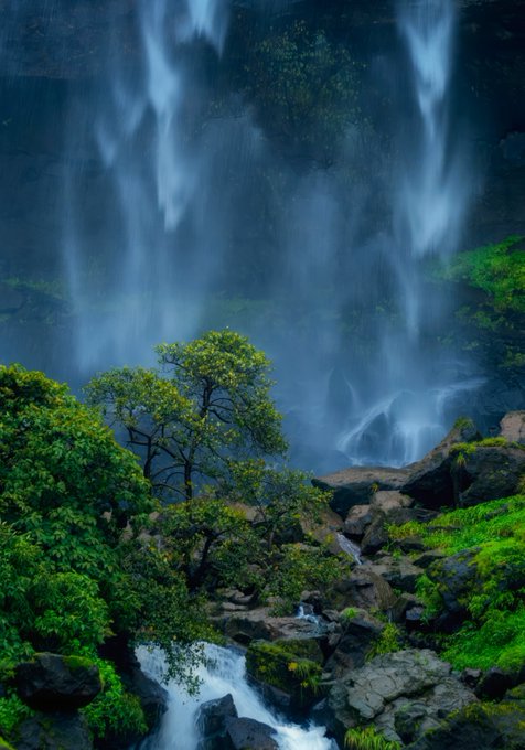A lush landscape photograph of a tall waterfall cascading down rocky cliffs, framed by green trees and mossy stones, with water flowing in the foreground.
