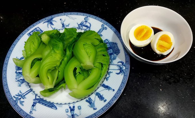 A simple vegetarian meal of blanched bok choy on a blue-and-white patterned plate, served alongside a small bowl of a halved hard-boiled egg in soy sauce, reflects the post's reflection on a meat-free morning after a night of football celebrations.