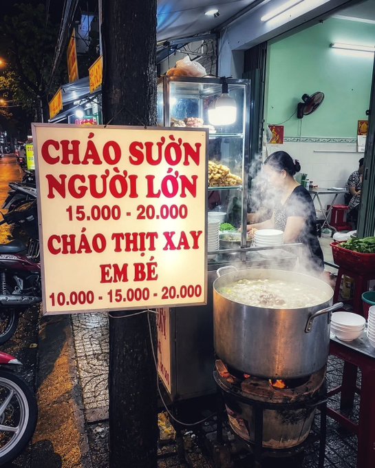 A Vietnamese street food stall with a yellow sign reading "CHÁO SƯỜN NGƯỜI LỚN 15.000 - 20.000 CHÁO THỊT XAY EM BÉ 10.000 - 15.000 - 20.000," with a large steaming pot of porridge over a fire.
