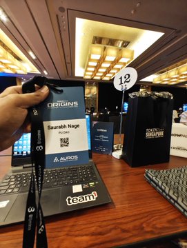 A photograph taken at an event labeled 'TOKEN2049 SINGAPORE,' showing a person's hand holding a conference badge for Saurabh Nage from the 'ORIGINS' summit, placed above a laptop and keyboard.