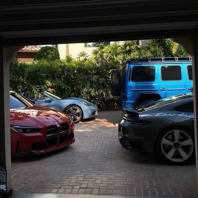 A view from inside a garage looking out at a driveway where a red BMW M4 is parked alongside a blue Mercedes G-Wagon and two silver Porsches.