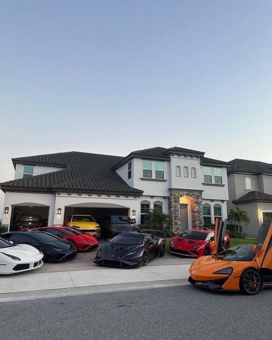 A large mansion with multiple luxury sports cars parked in the driveway, including Lamborghinis, a Ferrari, and a McLaren.