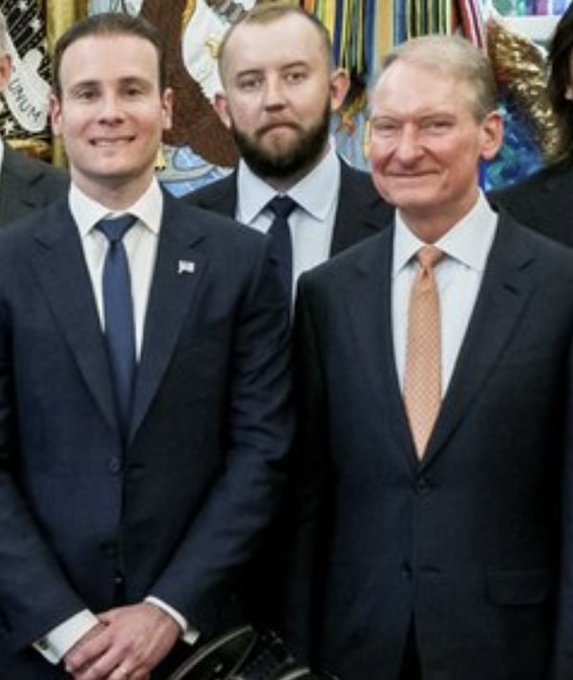 Two men in dark suits pose for a formal photograph, with the older man on the right wearing a peach tie and the younger man on the left wearing a blue tie and American flag pin.