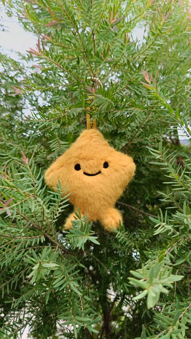 A fluffy yellow plush star with a smiling face and a partially visible tag reading "STAR" hangs among the green branches of a tree, perfectly matching the cheerful Monday vibes of trees, breeze, sunshine, and smiles shared in the post.