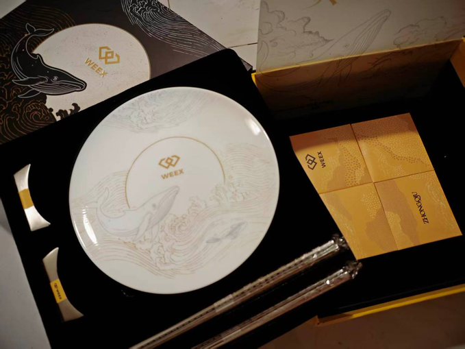 An elegant gift set featuring a white ceramic plate with intricate whale and ocean wave designs, accompanied by gold-accented utensils and yellow napkins, all branded with the WEEX logo.