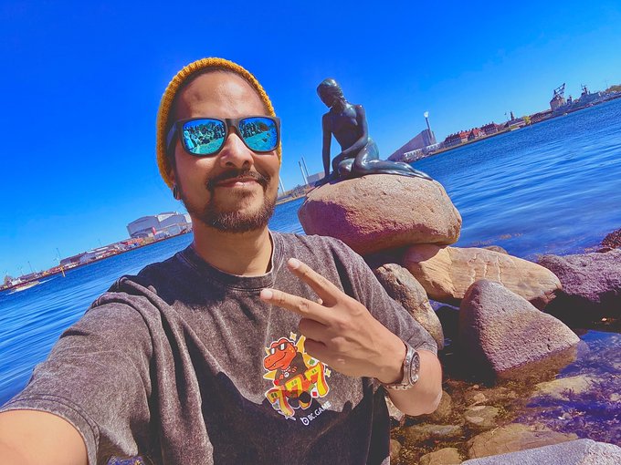 A man wearing reflective blue sunglasses and a beanie takes a selfie by the harbor, flashing a peace sign next to the iconic Little Mermaid statue in Copenhagen, Denmark. His gray t-shirt features a colorful dinosaur graphic, perfectly complementing his cheerful "GM" (Good Morning) greeting.