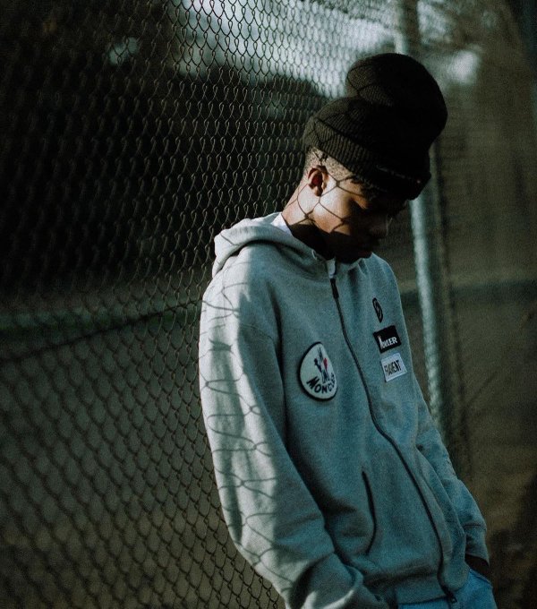 A masked person in a black beanie and gray hoodie leans against a chain-link fence, conveying a contemplative, mysterious mood that matches the post's reflective tone of sharing freely despite lacking material wealth like $ANSEM or cashcat. Visible text on the hoodie includes a circular "MONEY" patch and a rectangular "FACTORY CREW" tag.