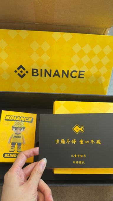 The image shows a hand holding up a small card with the words "Happy Children" written on it, along with an illustration of a boy wearing glasses. The card appears to be from Binance, as indicated by the text "binance" in the image description. The person is likely sharing this special edition card or gift with children for Happy Children's Day, expressing their appreciation and love for them.
