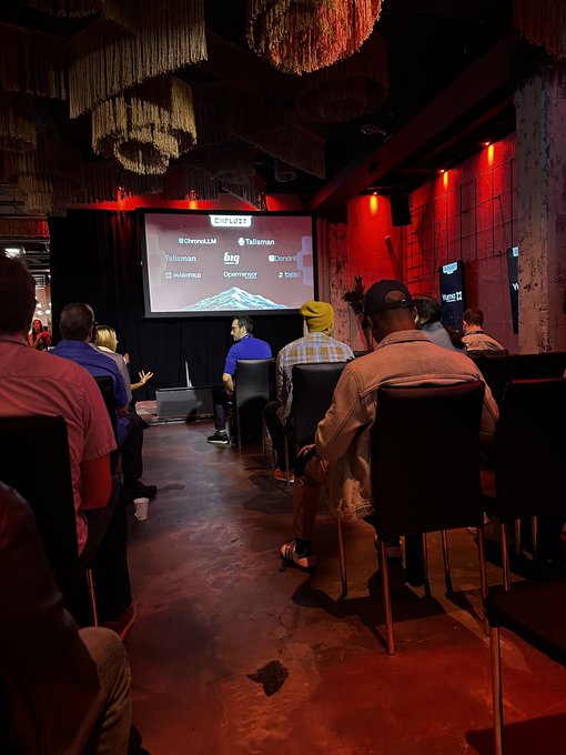 An audience gathers in a dimly lit venue with red ambient lighting and decorative hanging chandeliers, watching a presentation on a screen displaying "2025 LIST" alongside crypto-related logos and tickers including $CronosLM, $TelAIman, $ailcman, $Xlon, $novo, $paywayz, and $boo. The intimate setting with branded sponsorship visible on the screen aligns well with the post's reference to a "sexy venue" for what appears to be a crypto event involving $TAO.