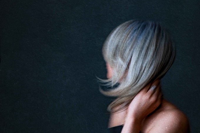 A soft, film-grain portrait of a woman with long blonde hair obscuring her face, her hand resting on her neck against a dark background.
