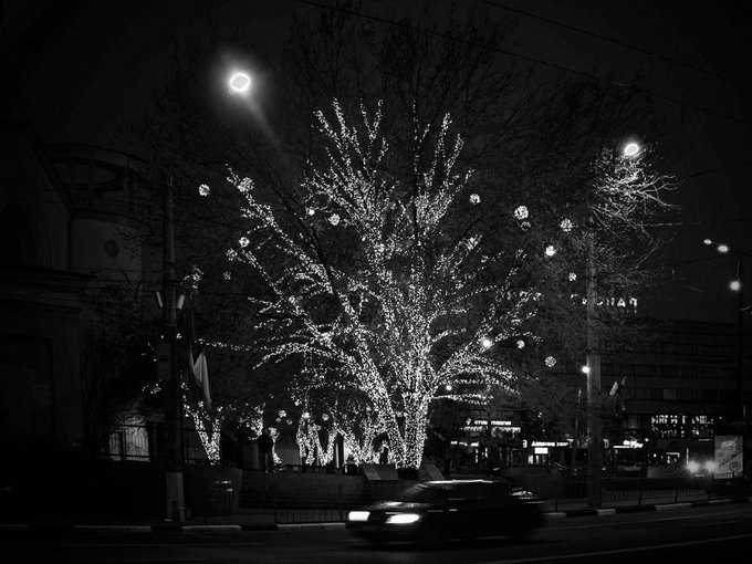 A black and white nighttime photograph showcases a large tree adorned with sparkling string lights and ornaments, illuminated by a glowing streetlamp, while a blurred car passes in the foreground. A partially visible sign reads "MAN," fitting the evening "Glow" theme conveyed in the post.