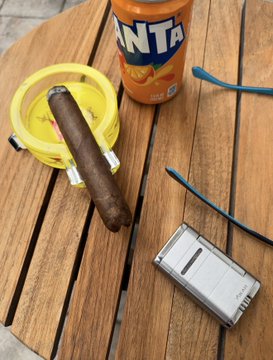 A cigar rests in a yellow ashtray next to a bottle of Fanta Orange on a wooden table, accompanied by two small frog figurines and a flip phone. The scene captures a casual moment of relaxation under an overhang during a rainy day, matching the post's theme of enjoying leisure time with a stogie, a soda, and frog companions while anticipating a late work night.